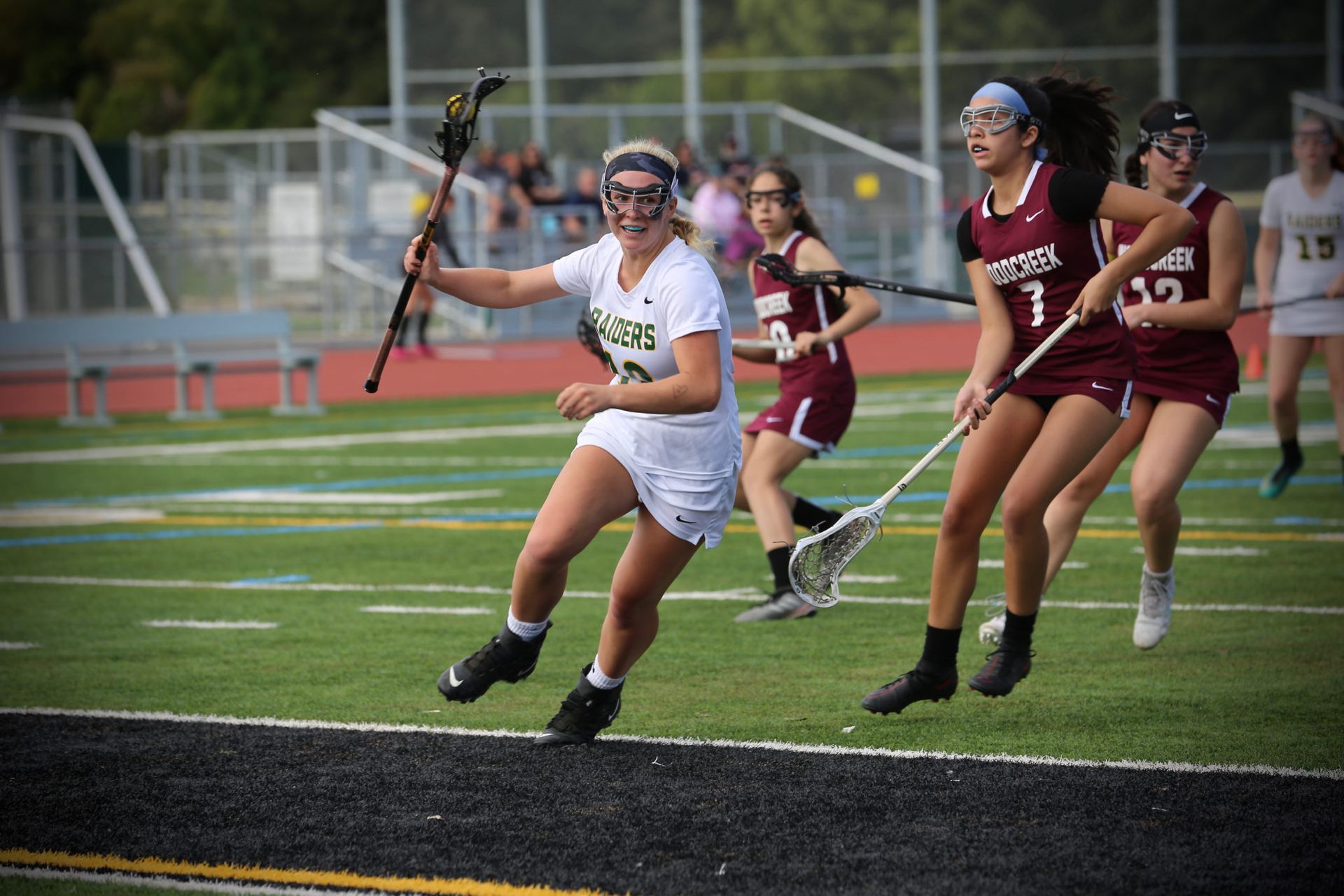 A lacrosse player in white runs across the field while being pursued by two opponents in maroon uniforms.