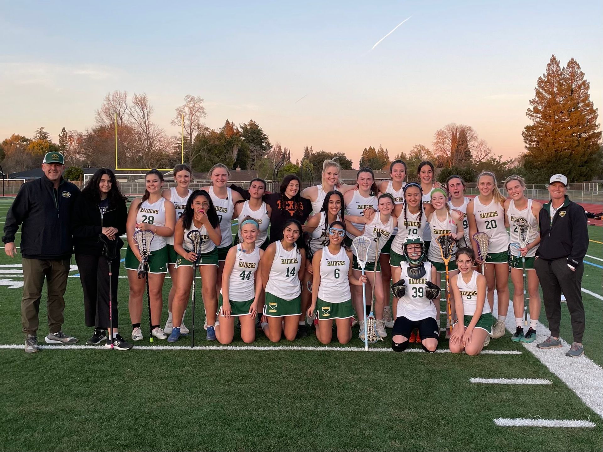 A lacrosse team posing for a group photo on a field at sunset, wearing white and green uniforms.