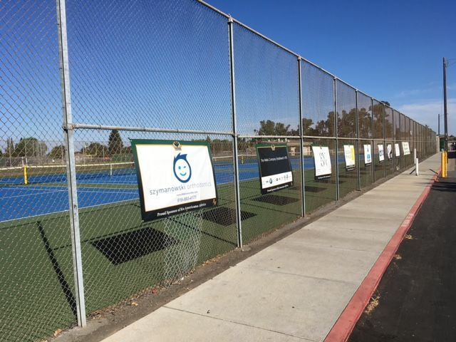 Tennis courts with several sponsor signs hanging along the chain-link fence bordering a paved sidewalk.