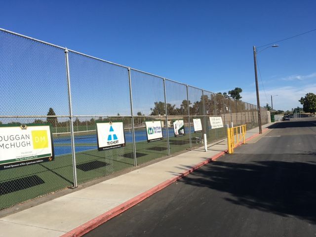 A row of advertisement signs attached to a chain-link fence along a sidewalk next to a tennis court under a blue sky.
