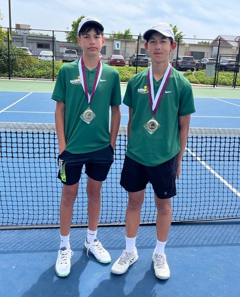 Two tennis players in green shirts and white caps stand on a blue court wearing gold medals around their necks.