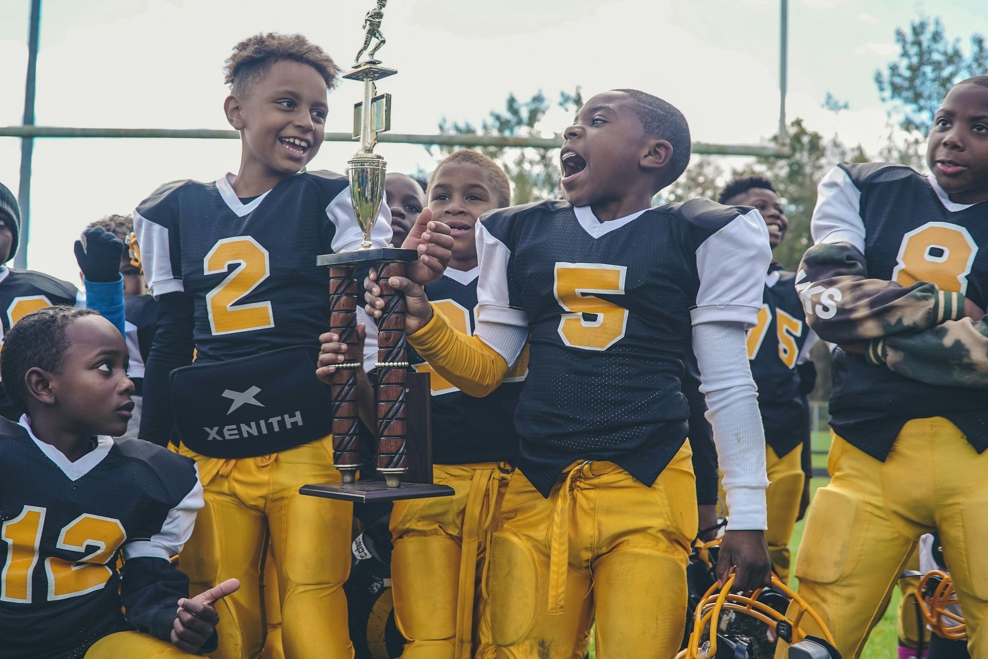 Young football players in black and gold uniforms cheer while holding a trophy on a field.