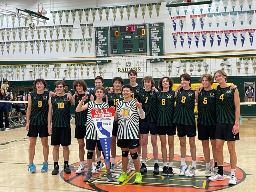 A volleyball team in dark green jerseys and black shorts stands in a row on a gym court, holding a tournament banner.