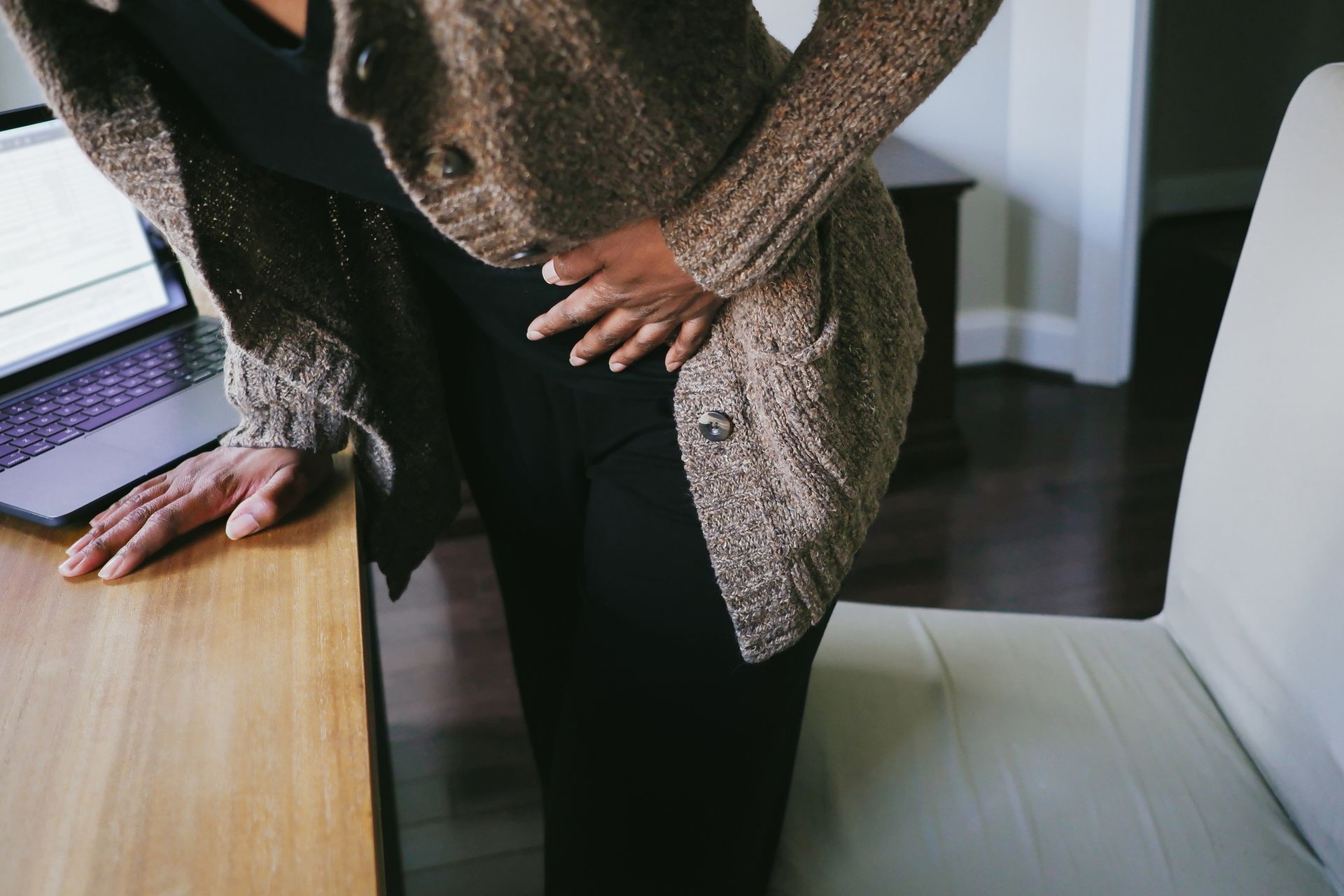 Woman clutching stomach, leaning on a desk with a laptop. Brown sweater, black pants, indoor setting. Woman clutching stomach, leaning on a desk with a laptop. Brown sweater, black pants, indoor setting.