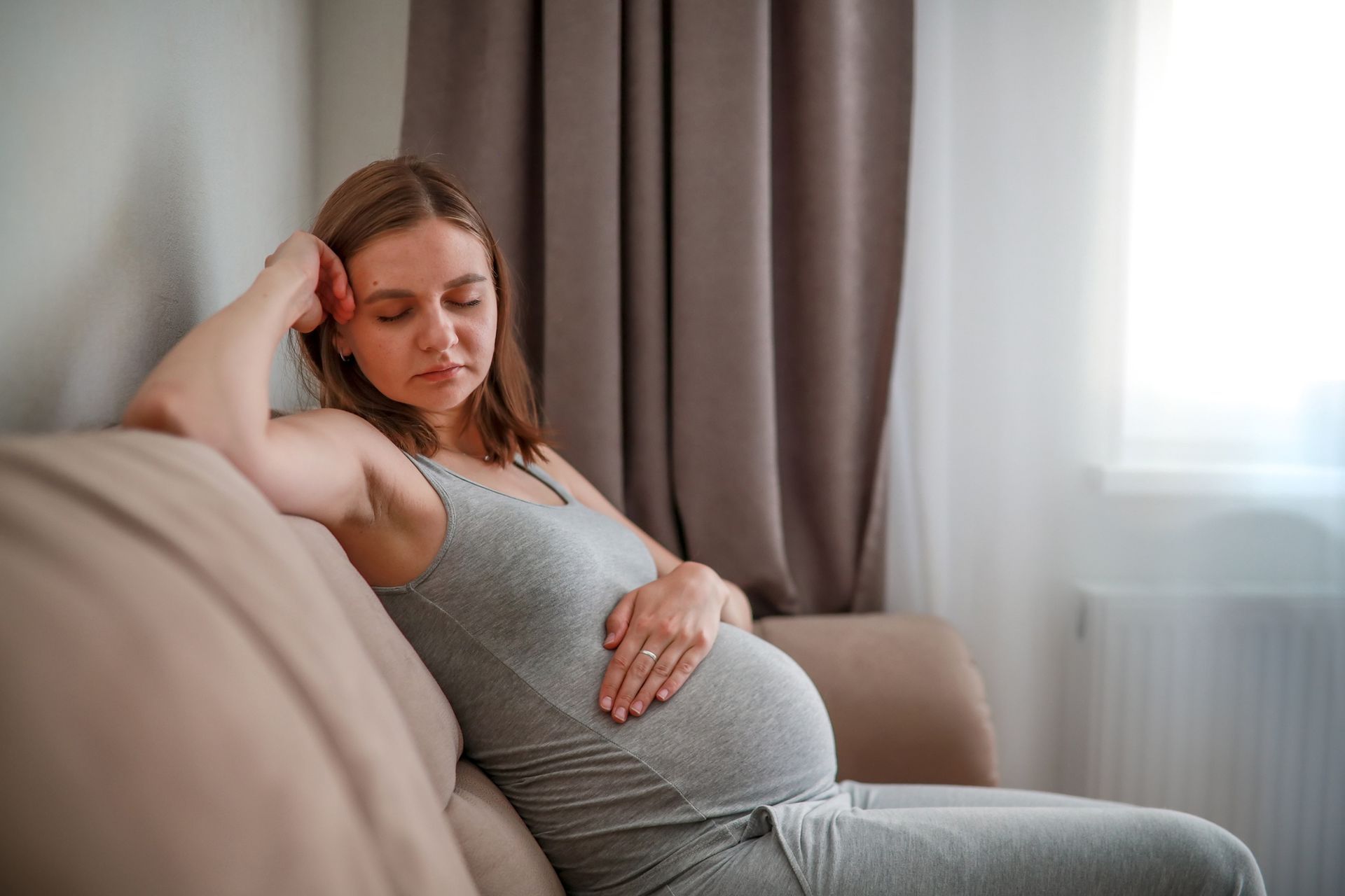 Pregnant person sitting on a couch, resting hand on belly, looking down. Pregnant person sitting on a couch, resting hand on belly, looking down.