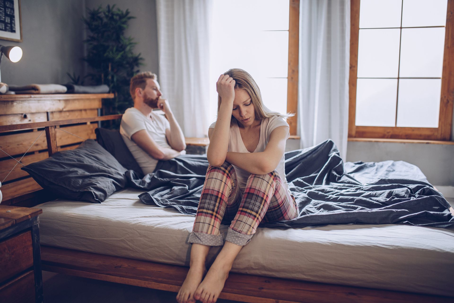 Woman with head in hands sitting on bed, man looking away, appears distressed, bedroom setting. Woman with head in hands sitting on bed, man looking away, appears distressed, bedroom setting.