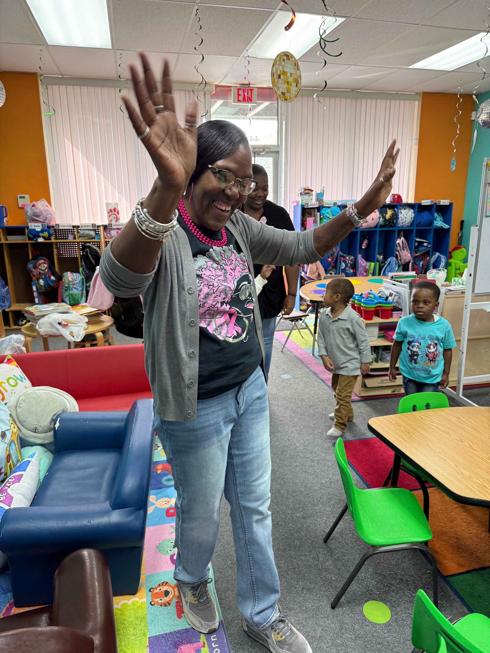 Woman waving arms, smiling in a colorful classroom. Two children watching near a table.