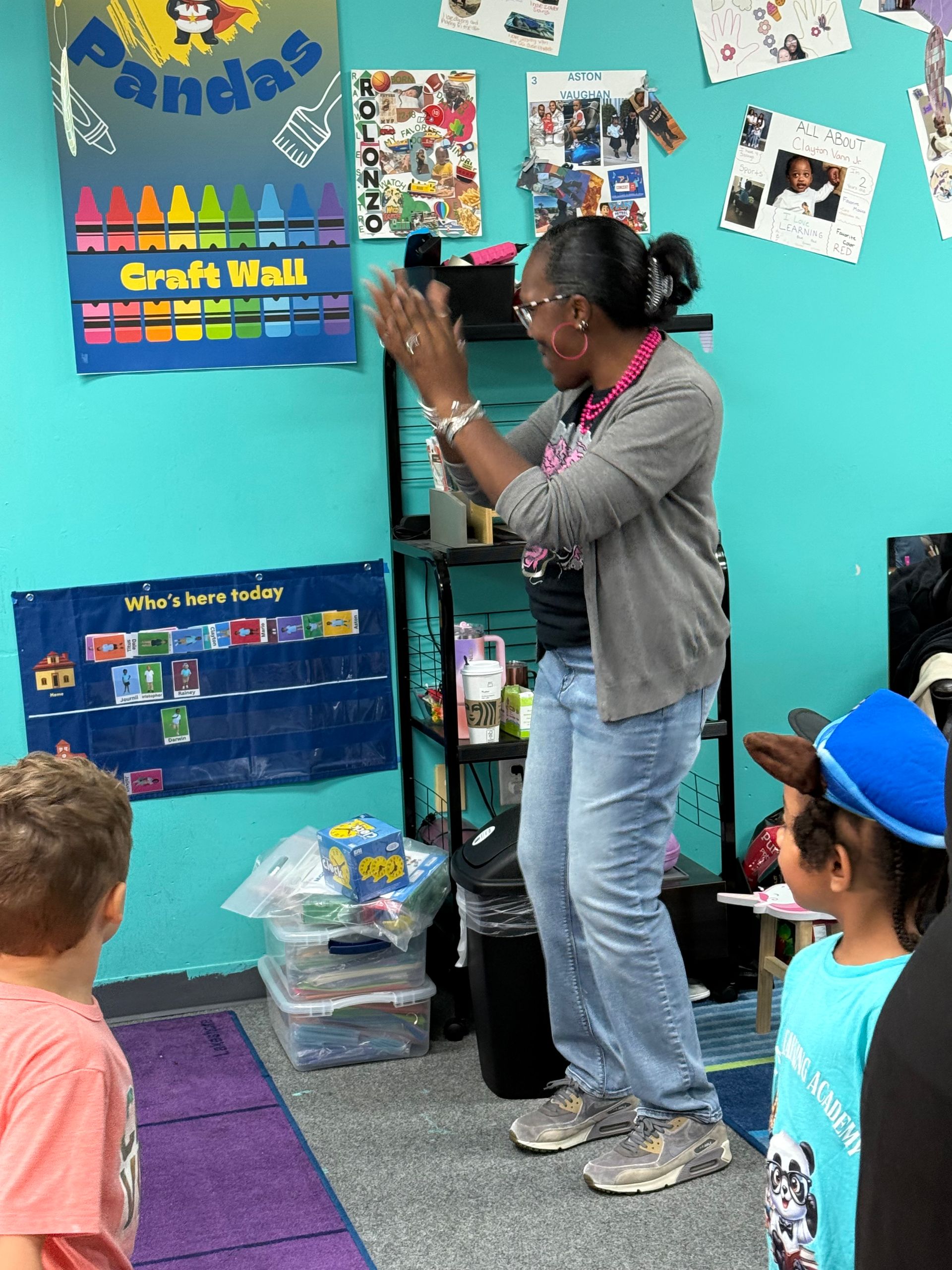 Teacher clapping in a classroom, children watching. Blue wall, shelves, and craft wall in background.