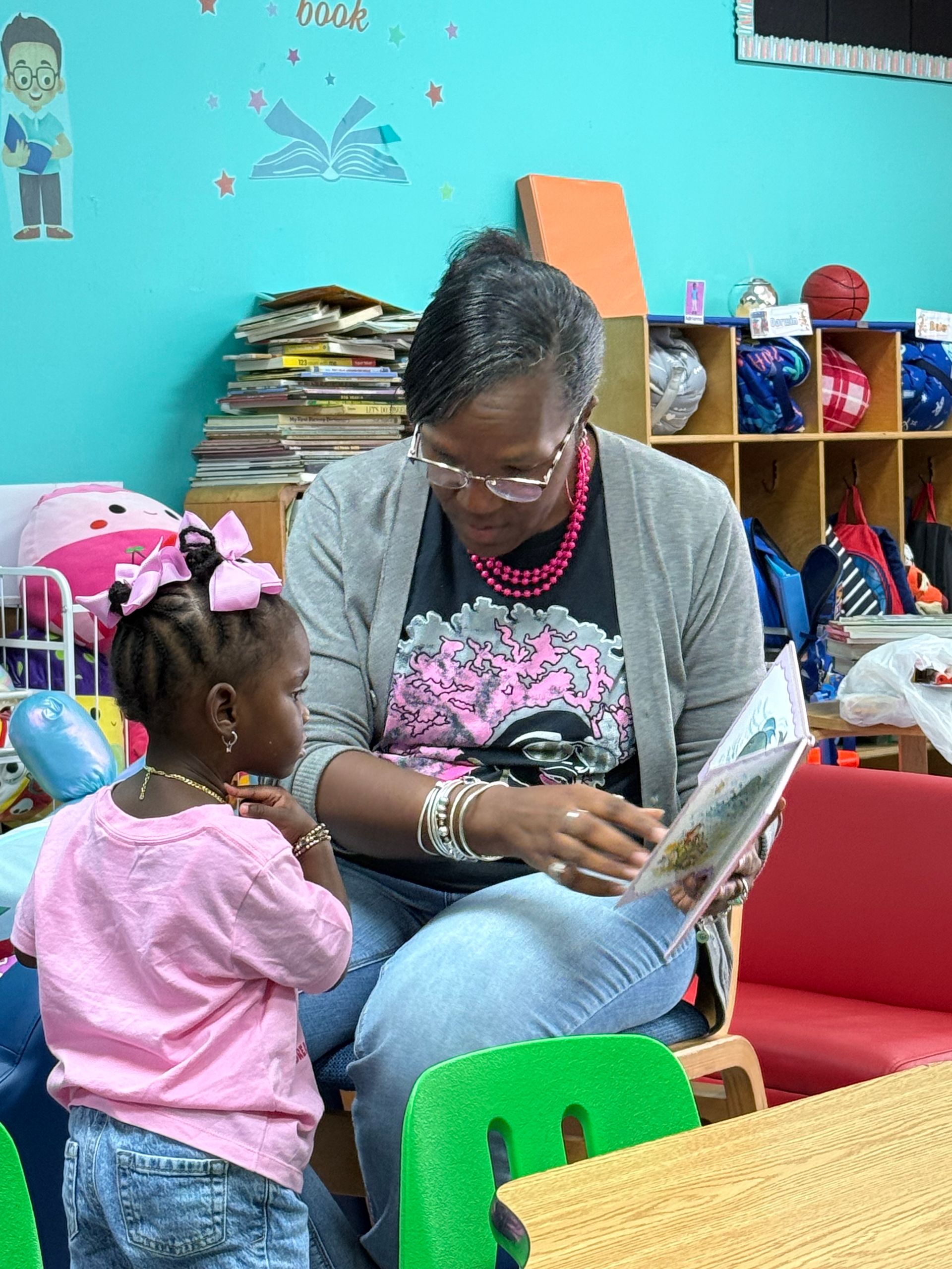 Woman reading to a child in a colorful classroom; the child in pink, the woman in jeans and a gray cardigan.
