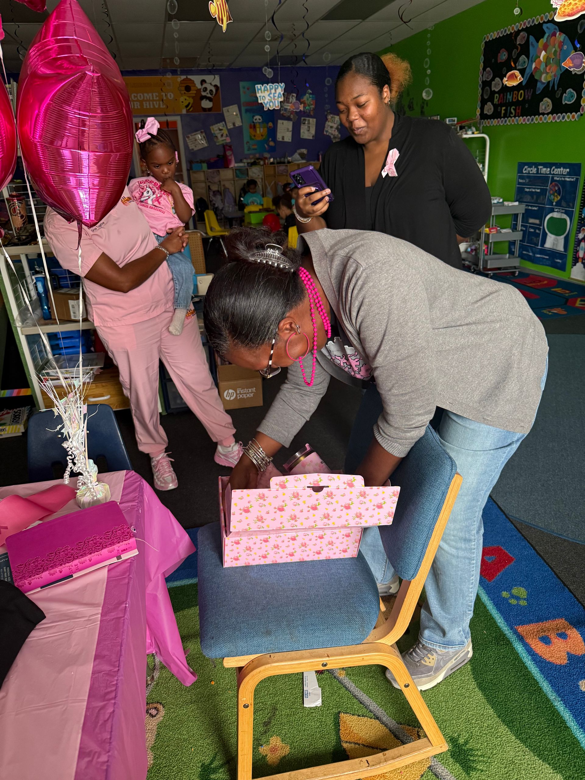 People in a room with pink decorations and a child opening a gift box.