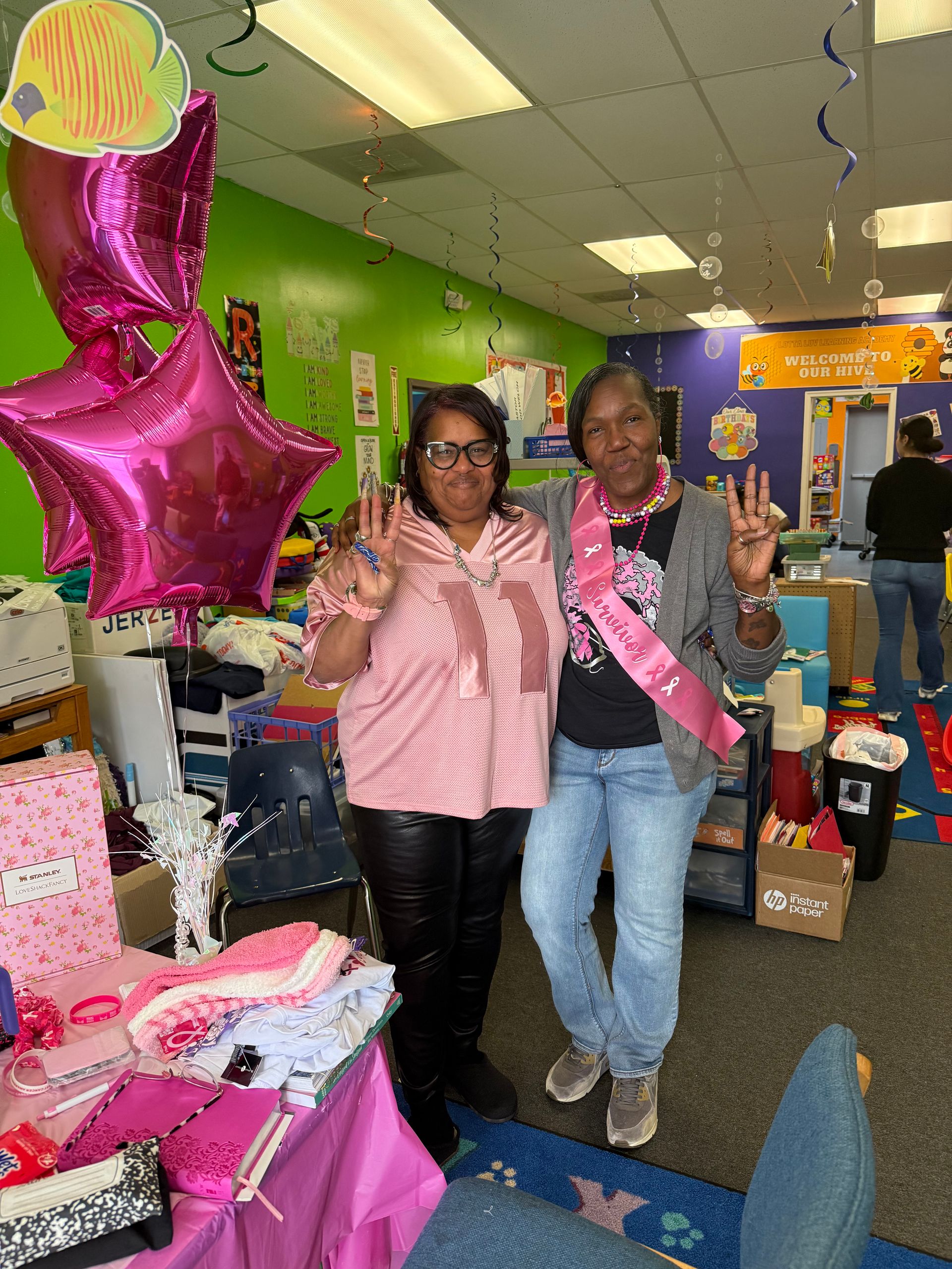 Two women indoors, smiling, posing with “3” hand gestures, near pink balloons and gifts.