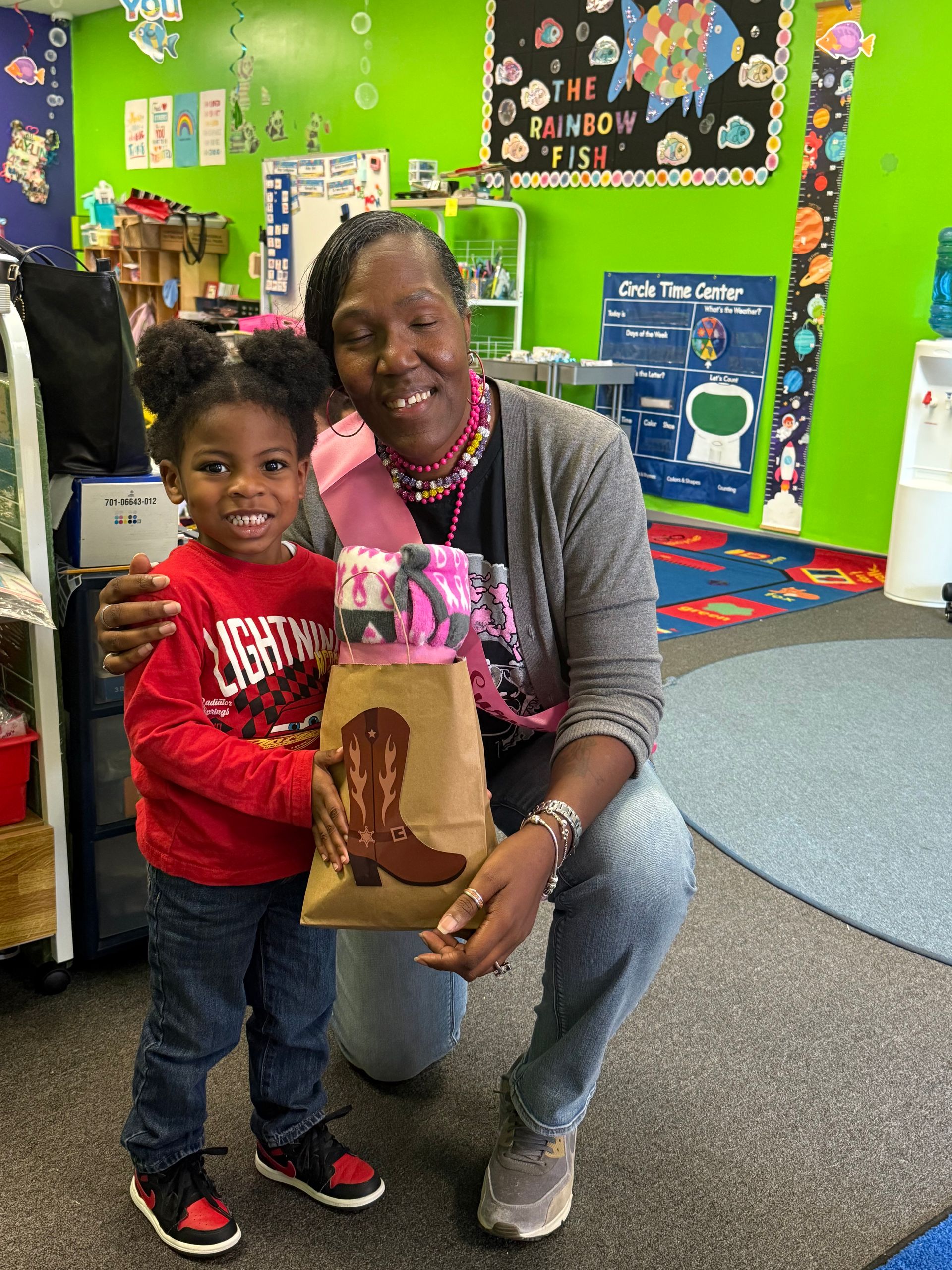A smiling child and woman in a colorful classroom, holding a decorated gift bag.