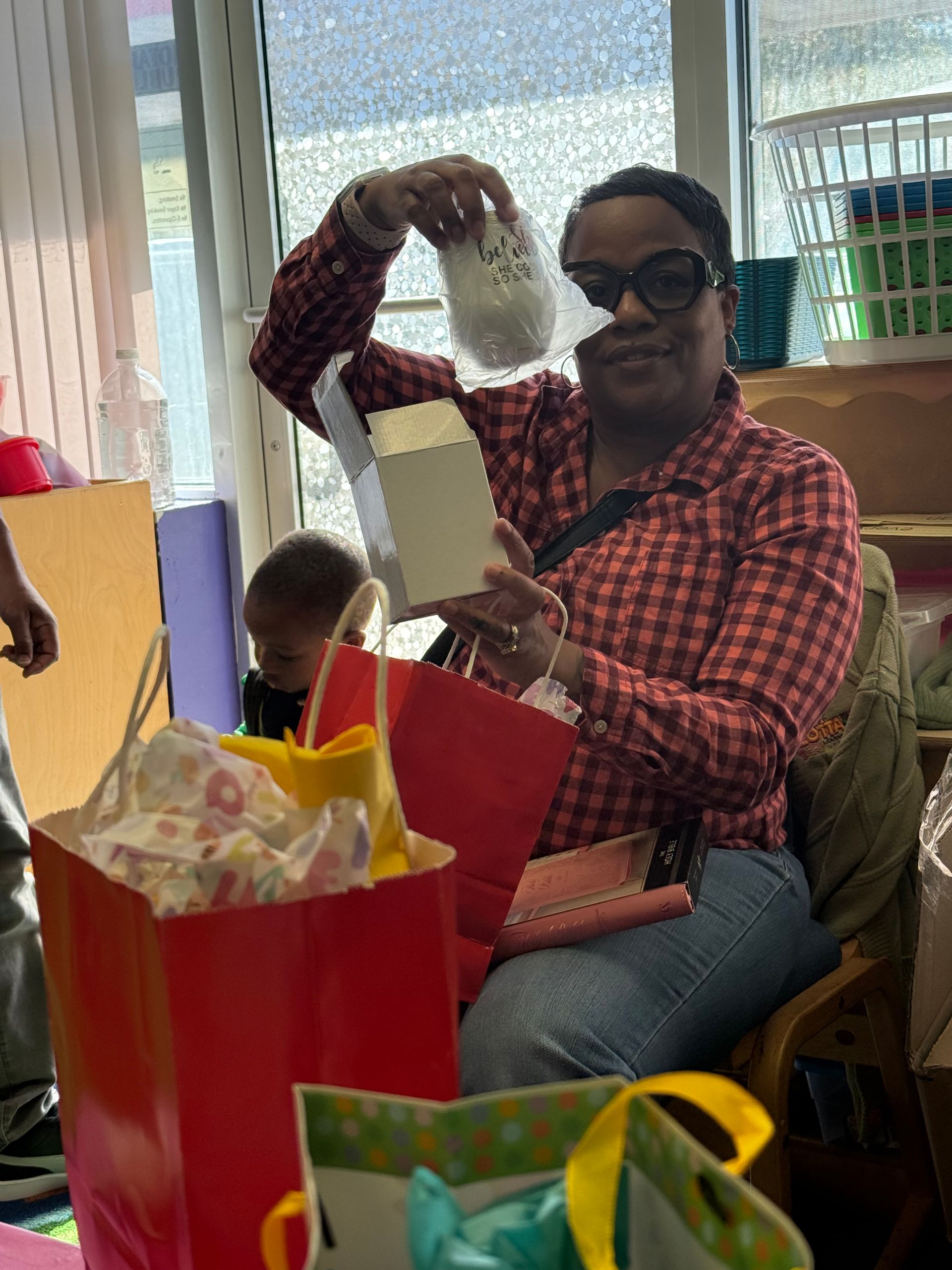 Woman holding a white jar with writing, surrounded by gift bags and a child in a classroom setting.