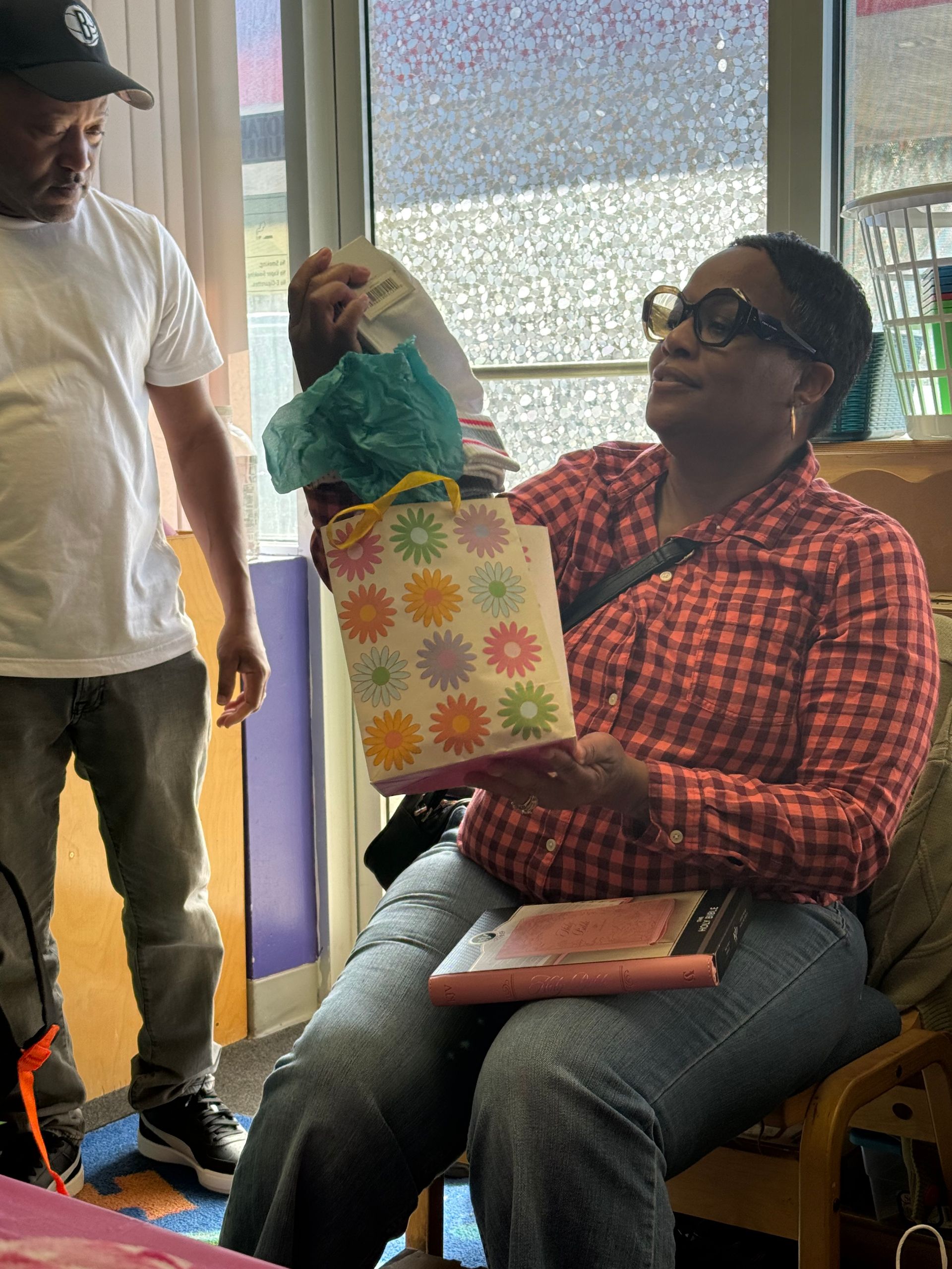 Woman seated, holding a gift bag. Man stands nearby. Interior setting with colorful accents.