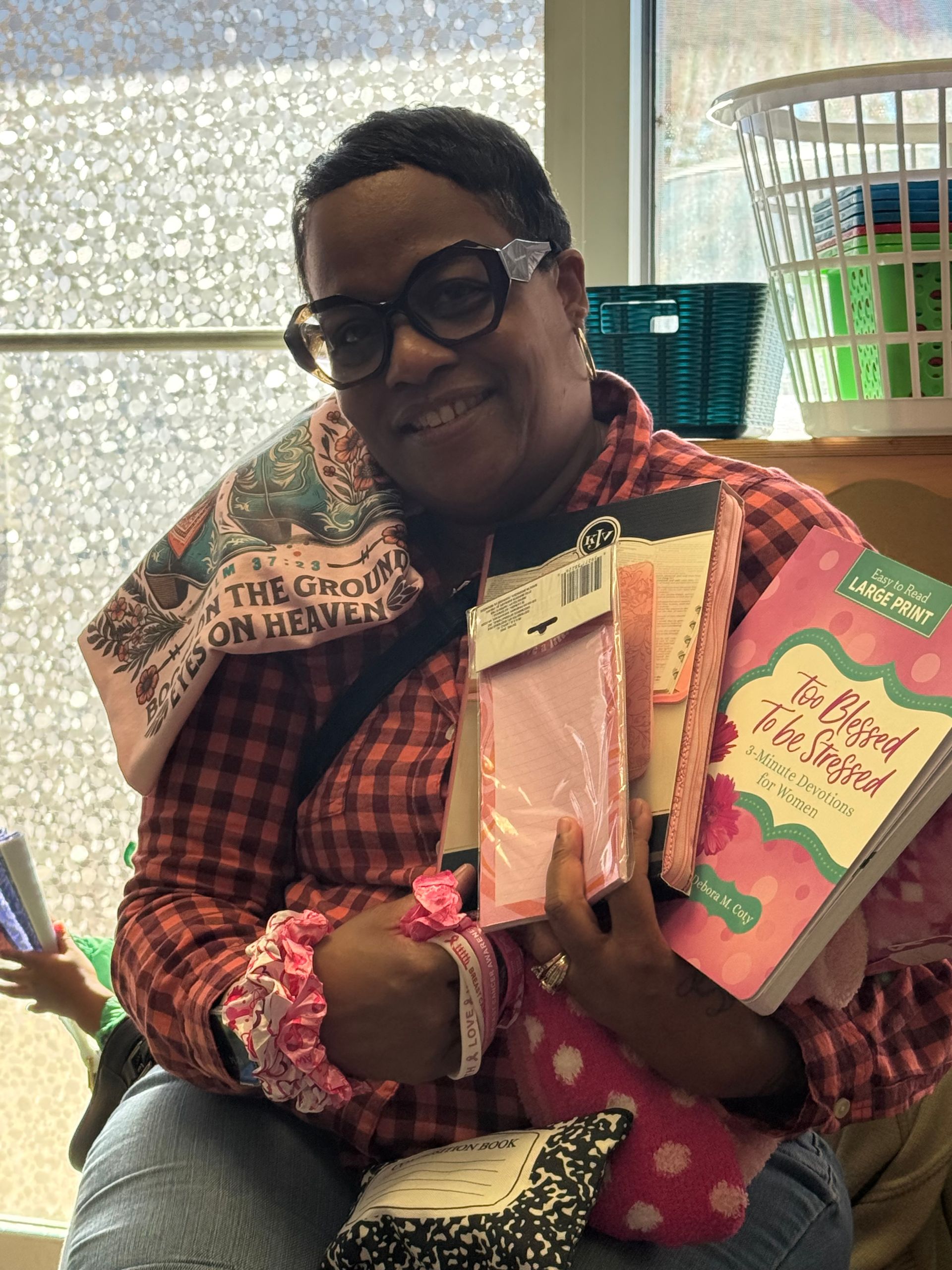 Woman smiling, holding pink gifts and books; wearing glasses and plaid shirt. Sunlight through the window.