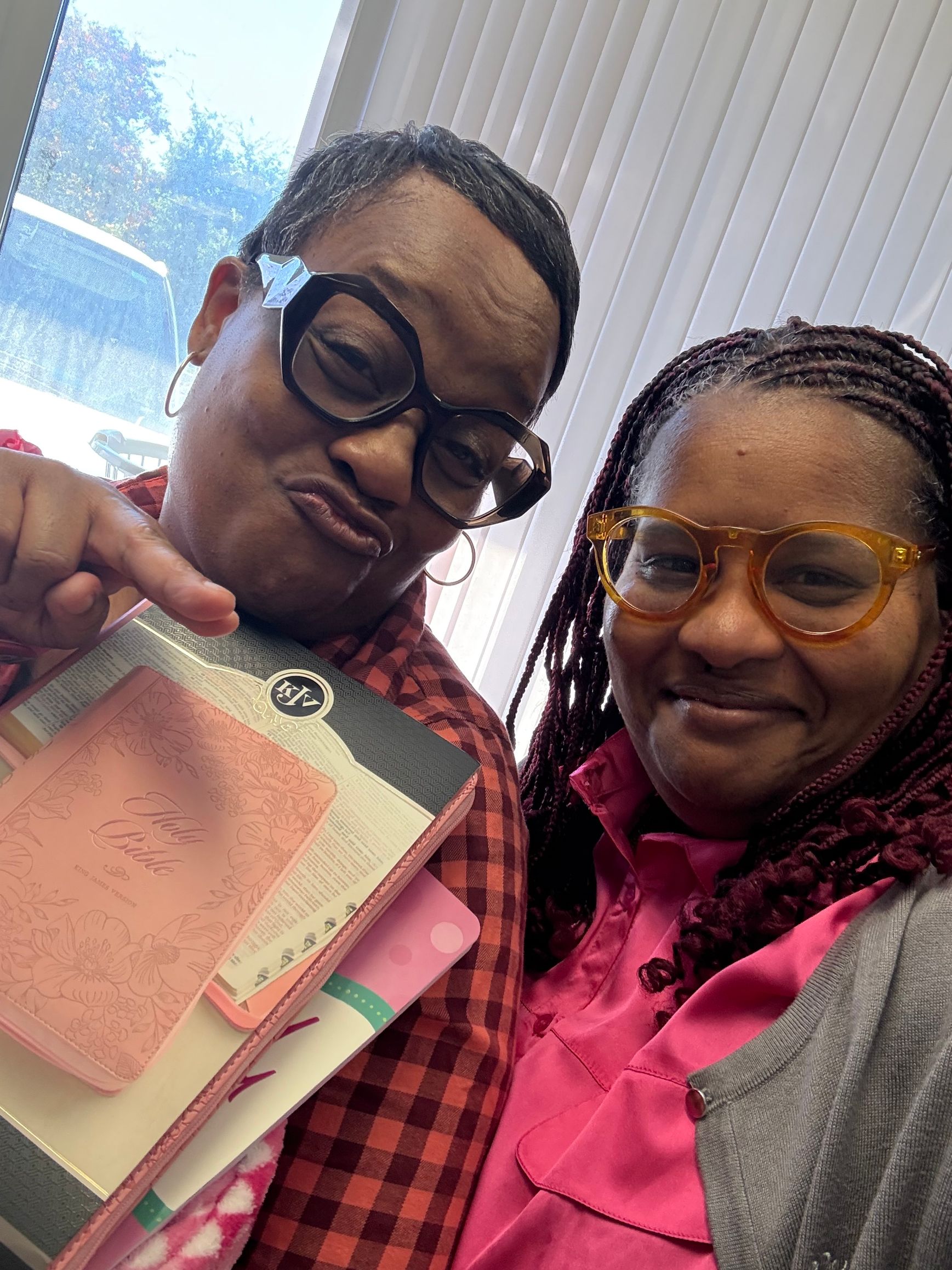 Two people smile, holding notebooks. One points. Pink and orange glasses are worn.