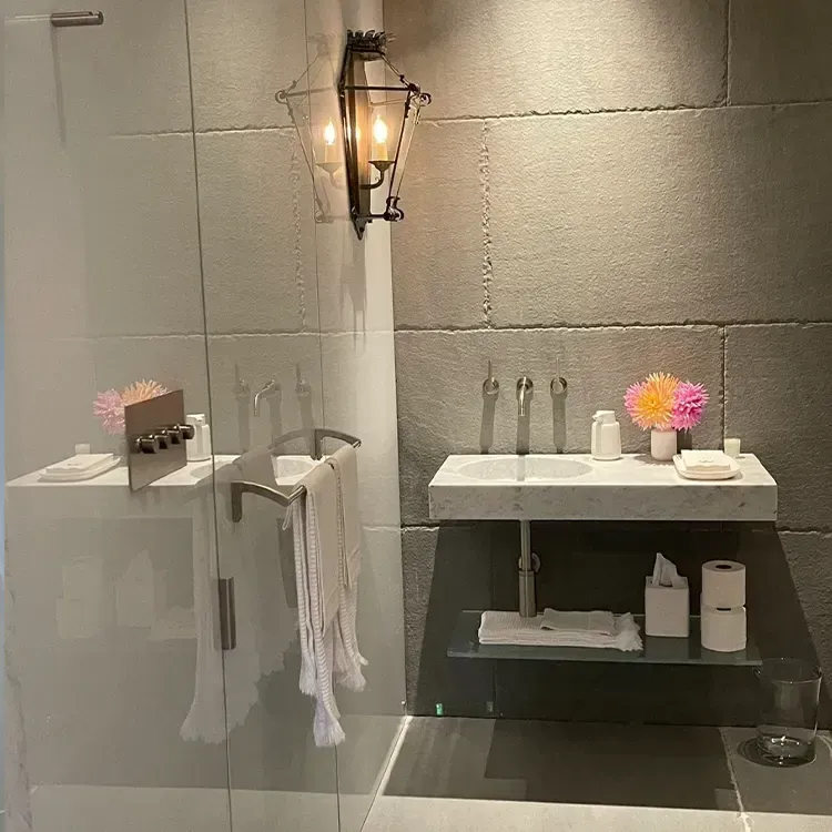 Modern bathroom vanity with lit wall sconce, sink, towels, and pink flowers on shelves.
