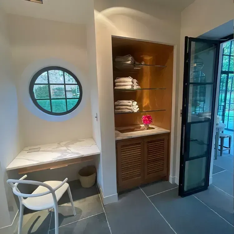 Modern bathroom with round window, marble vanity, built-in shelves, and open glass door to a patio.