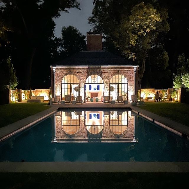 Nighttime view of a lit brick house reflected in a calm swimming pool, with trees and garden lights.