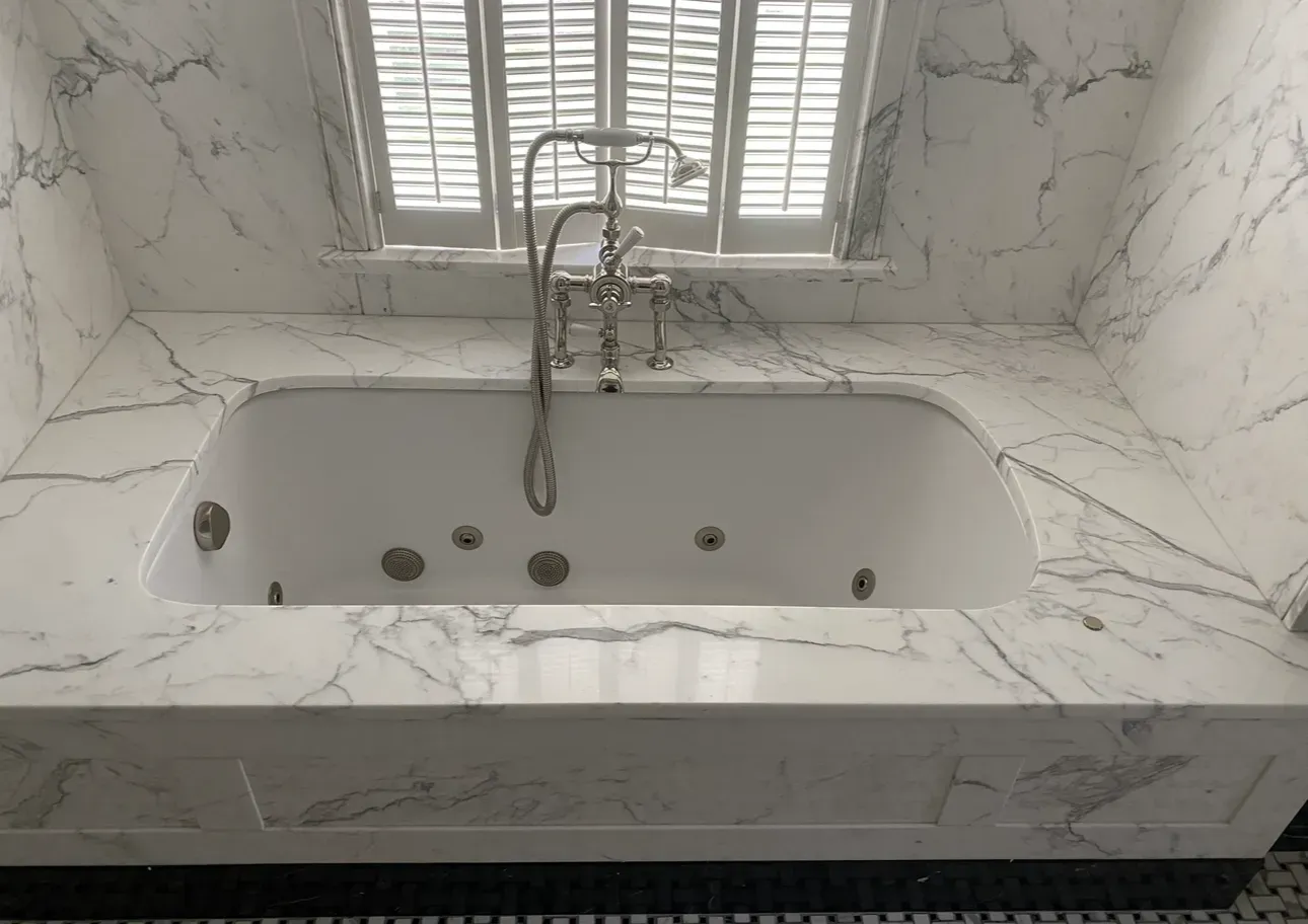 Modern white marble bathroom with a deep soaking tub beneath a window and faucet fixtures