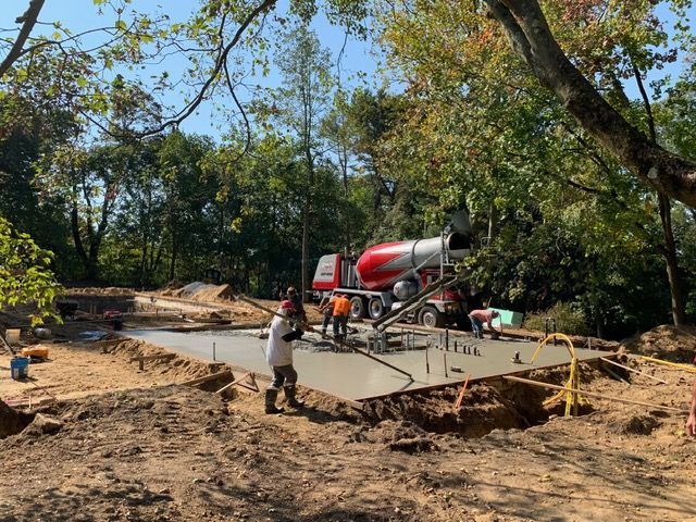 Concrete slab being poured at a wooded construction site, with a red cement mixer truck and workers nearby