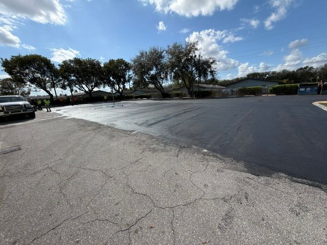 Empty parking lot with cracked pavement, trees, and a blue sky with clouds.