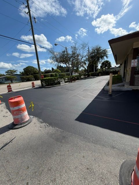 Sunny parking lot with orange traffic barrels, asphalt driveway, trees, and a building on the right.