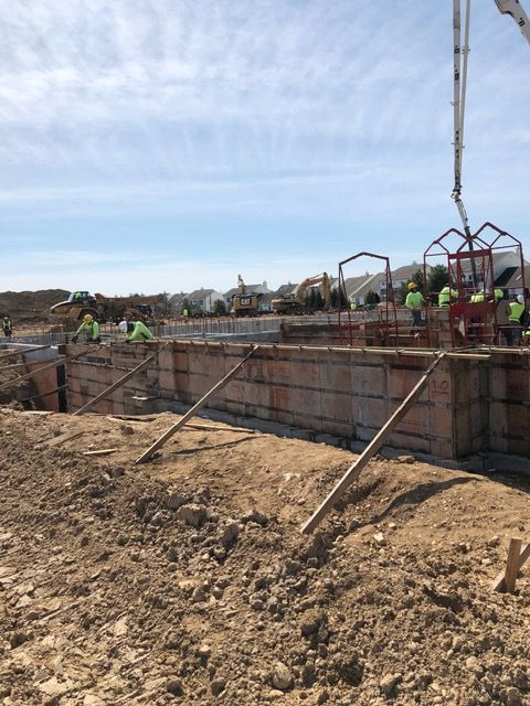 Construction site with concrete foundation walls, dirt foreground, and workers near equipment under a blue sky