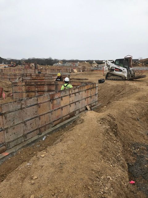 Construction site with workers beside a large concrete foundation and excavator on dirt terrain