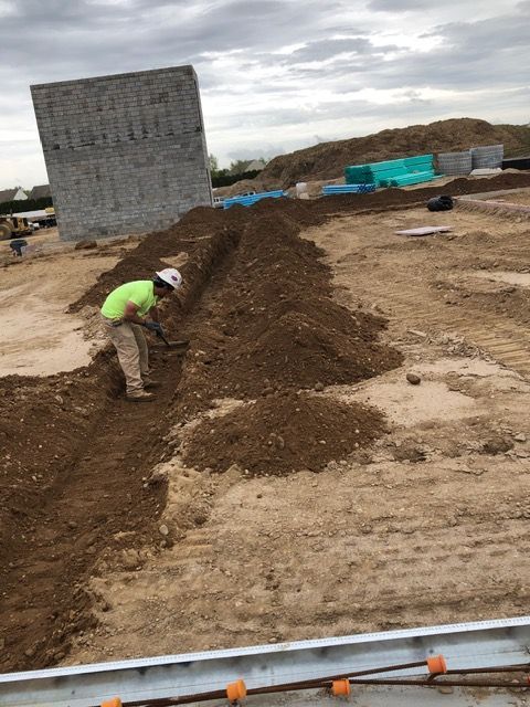 Worker spreading dirt along a trench at a construction site, with concrete blocks and equipment nearby