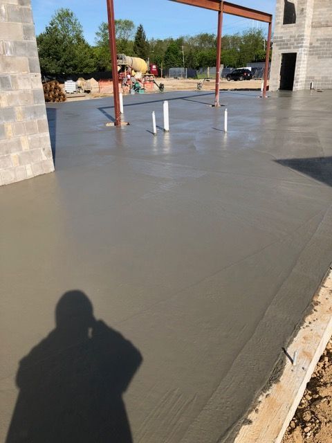 Freshly poured concrete slab beside a stone building with metal posts and construction materials in the background