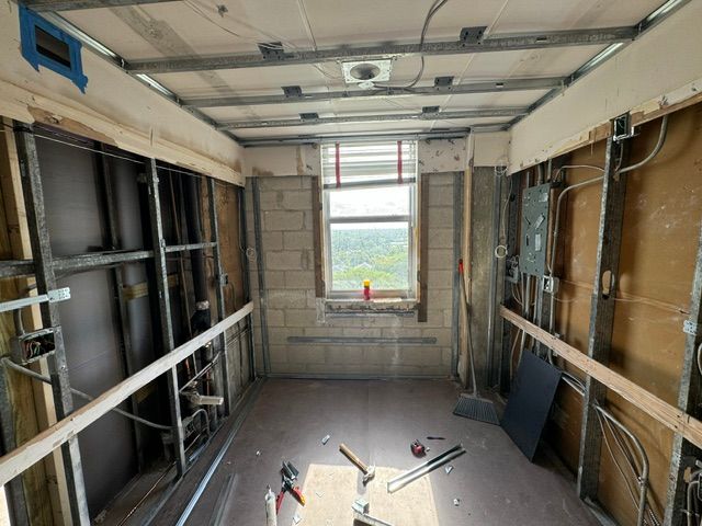 Empty room under renovation with exposed walls, ceiling beams, and tools on the floor near a window.