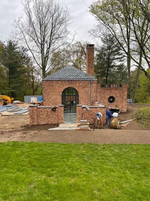 Brick garden structure under construction with arched doorway, chimney, and workers in a landscaped yard