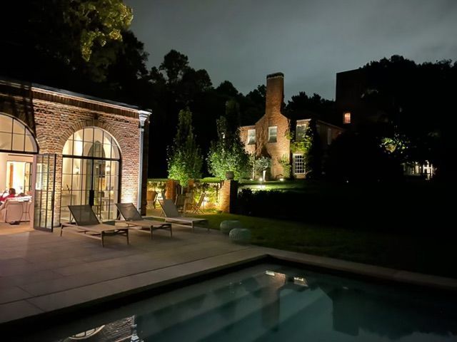 Lit poolside patio beside a brick house at night, with lounge chairs and warm garden lighting