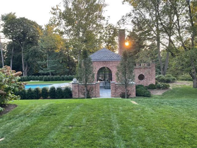 Brick garden pavilion beside a manicured lawn and pond at sunset, framed by trees and hedges