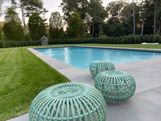 Turquoise wicker stools beside a backyard swimming pool with lawn and trees at dusk