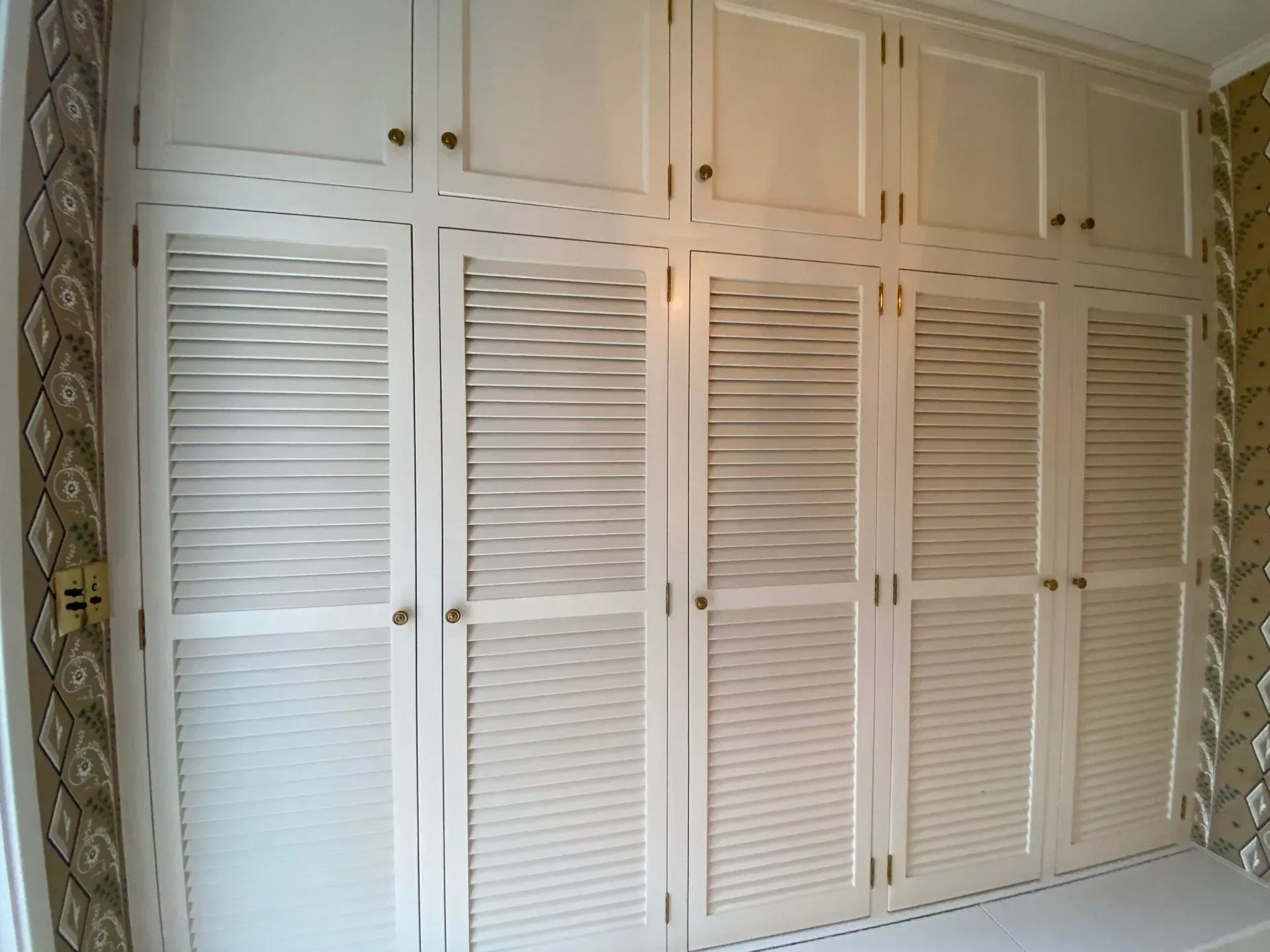 White louvered closet doors in a bedroom with built-in cabinets above and patterned wallpaper on the right.