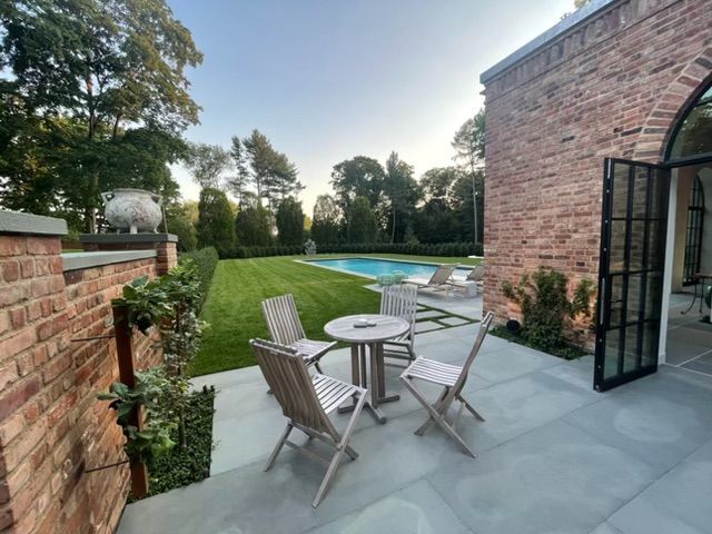 Patio with wooden chairs and round table beside a garden and pool, next to a brick house with open doors