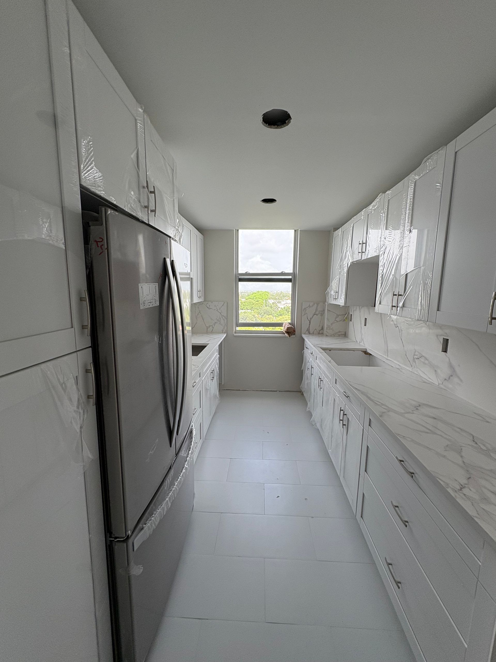 Narrow white galley kitchen with stainless fridge, long counters, and a window at the end.