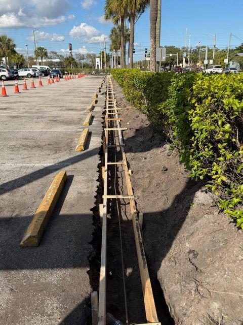 Construction trench along parking lot curb with wooden formwork, orange cones, and palm trees in the background