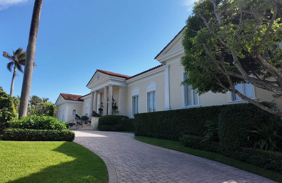 White villa with red-tile roof and palm trees along a curved paved driveway under a blue sky