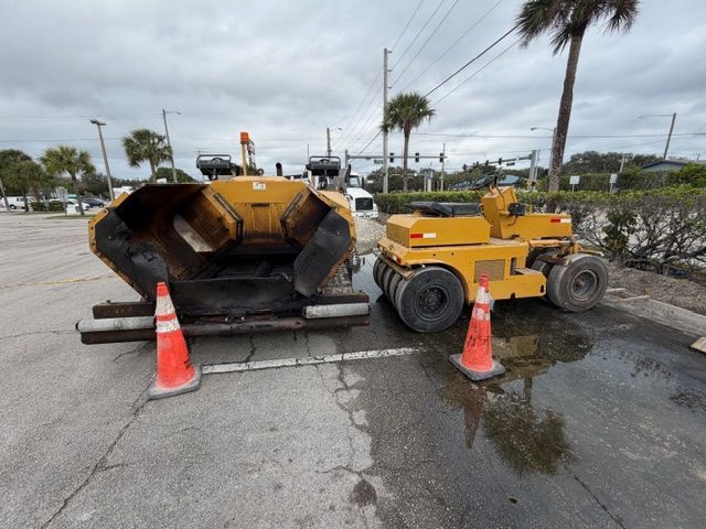 Yellow road construction machine beside asphalt paver on wet street with traffic cones, near palm trees