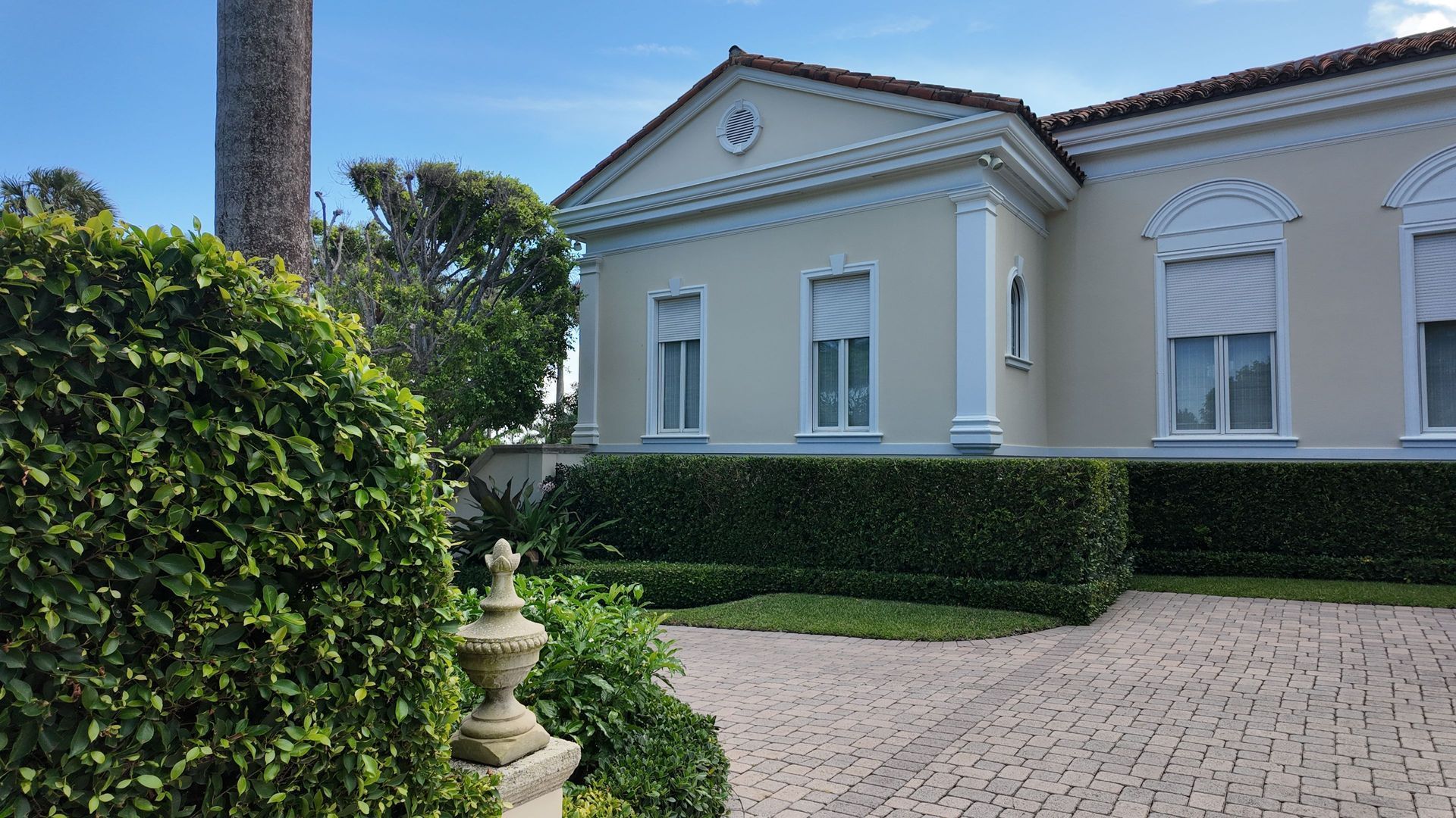 White classical mansion with columns, manicured hedge, palm tree, and cobblestone driveway under clear blue sky