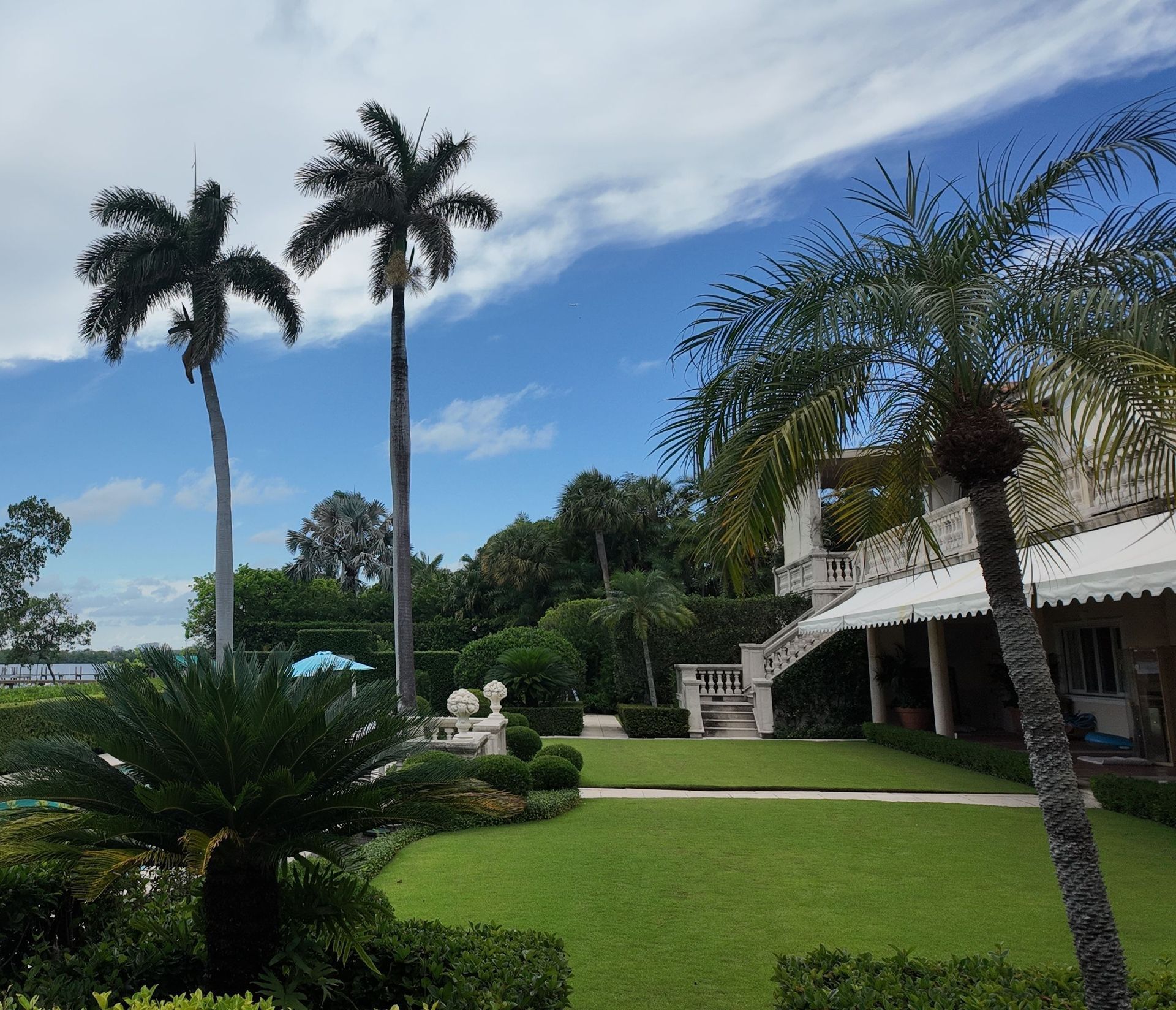 Tropical garden with palm trees, green lawn, and a white house under a partly cloudy sky