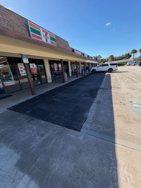 Strip mall storefronts with a 7-Eleven sign and a black ramp leading to the entrance under a blue sky