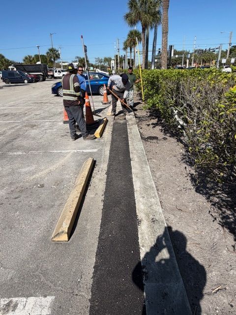 Workers installing curb edging along a sidewalk in a sunny parking lot, with tools and lumber nearby.