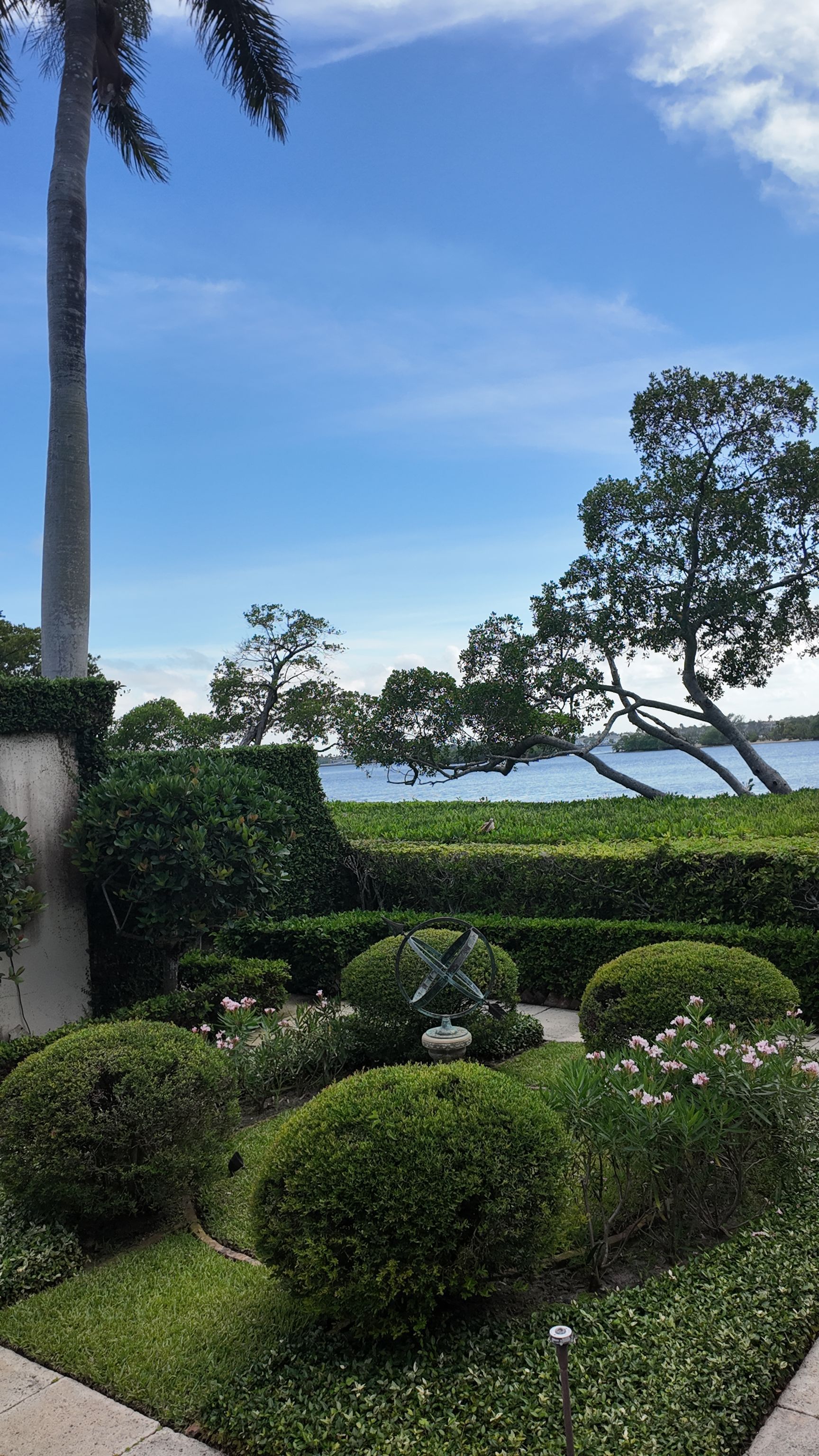 Manicured garden hedges and palm trees overlooking a bright blue ocean under a partly cloudy sky