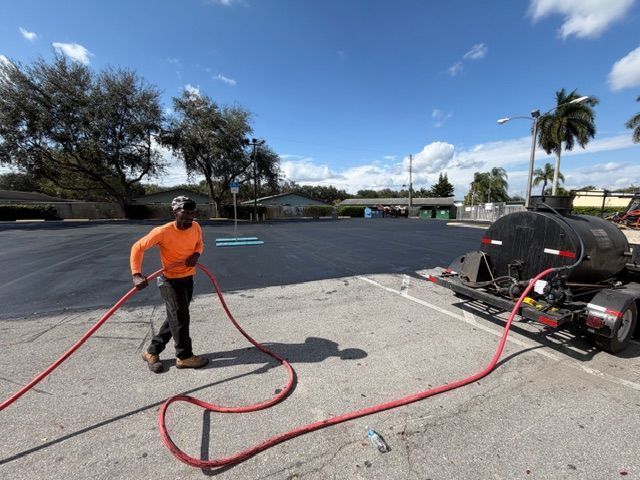 Worker in orange shirt handling a red hose beside a trailer-mounted machine in a parking lot under a blue sky