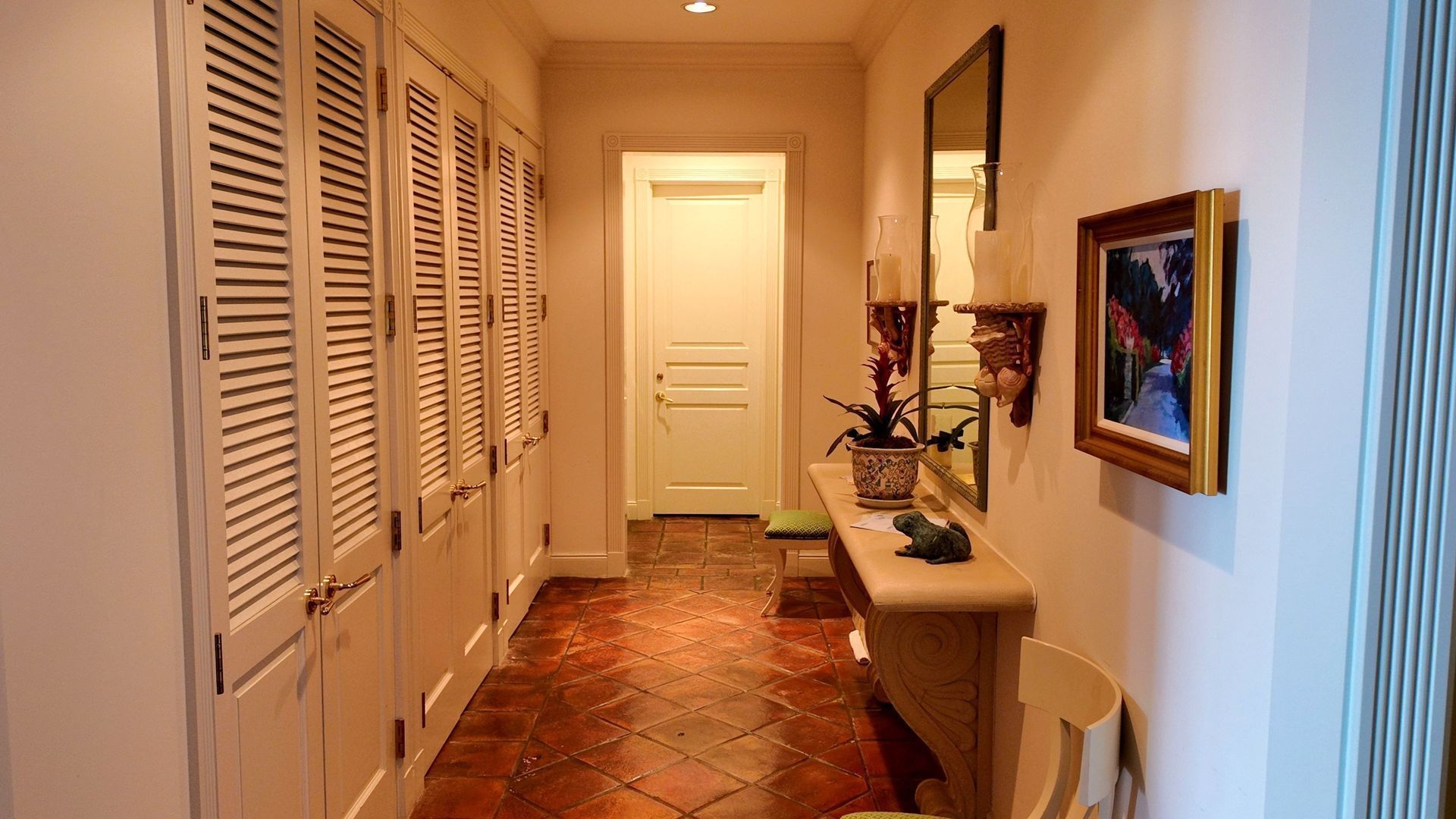 Narrow hallway with white closet doors, a console table, mirror, and framed art on a tiled floor.