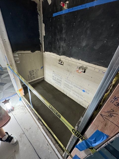 Caution-taped construction pit beside a black wall with exposed framing and a person’s legs at left.